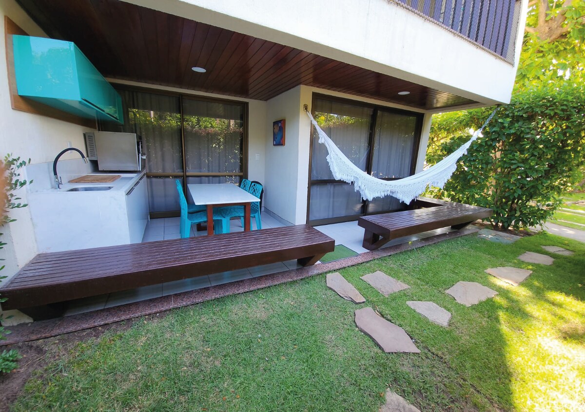 An outdoor area features a shaded patio with a dining table and contrasting teal chairs. Two wooden benches are positioned around the space, while a hammock is suspended between the posts. Lush greenery surrounds the area, providing a serene atmosphere.