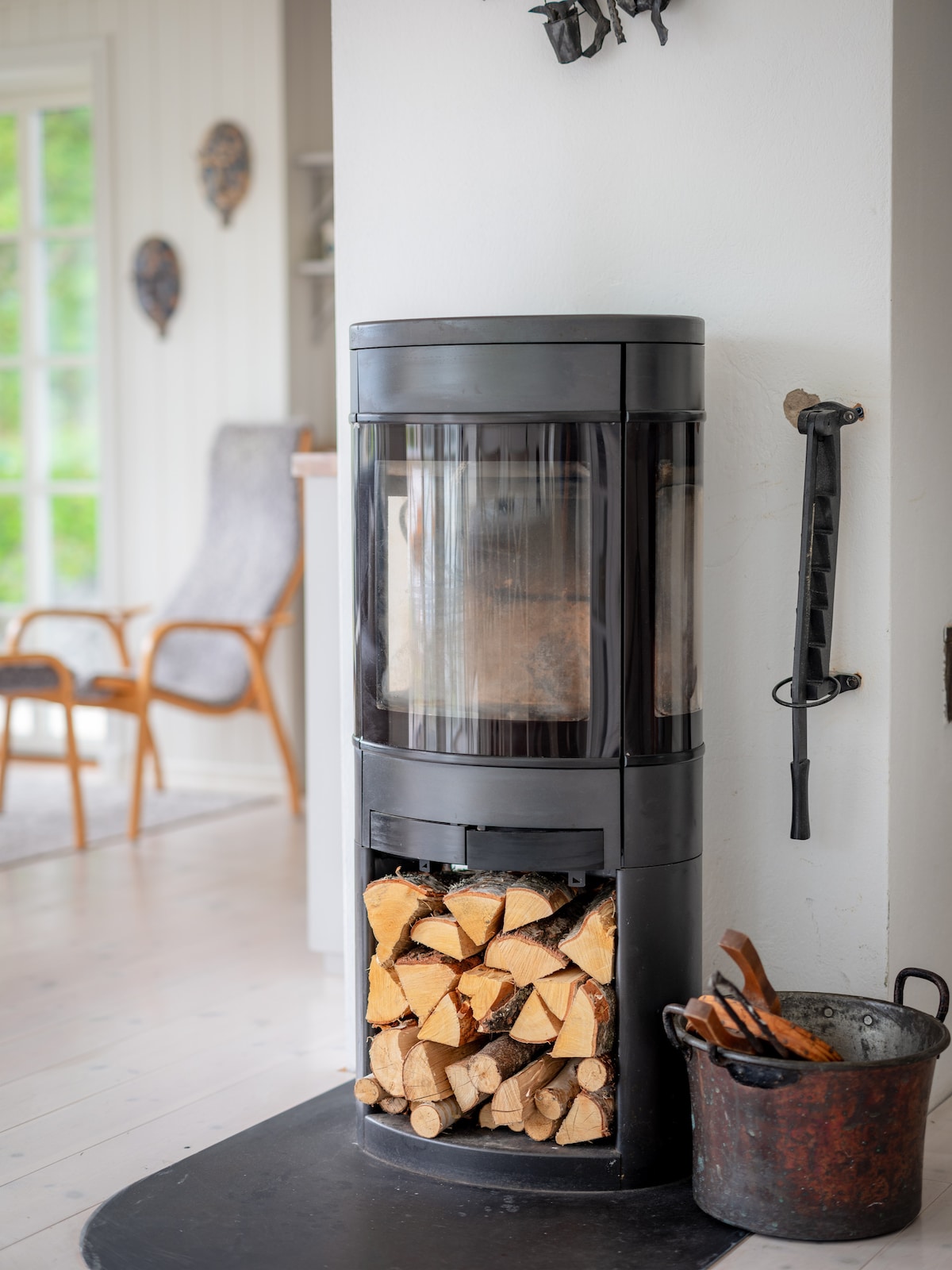 A modern wood stove is positioned in a bright corner, featuring a glass front that displays the wood fire within. Stacked firewood is neatly arranged below, while a burnished metal bucket and a tool set are placed beside the stove, adding functionality to the space.