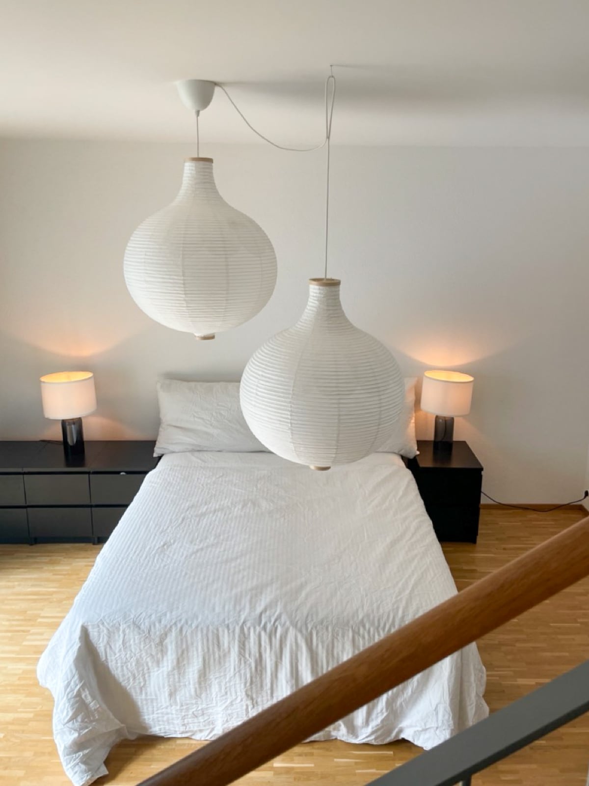 A cozy bedroom is showcased with a spacious bed covered in white bedding. Two large round pendant lights hang above, complemented by bedside lamps on dark dressers. Natural light enters from an unseen window, creating a serene atmosphere.