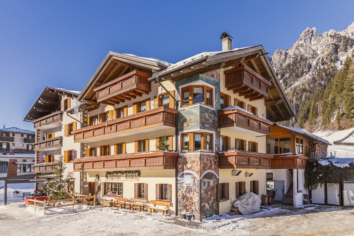 The three-story building features a chalet-style design with multiple balconies and wooden shutters. The lower level includes outdoor seating and decorative murals, while a snowy landscape and pine trees frame the structure against a backdrop of rugged mountains and a clear blue sky.
