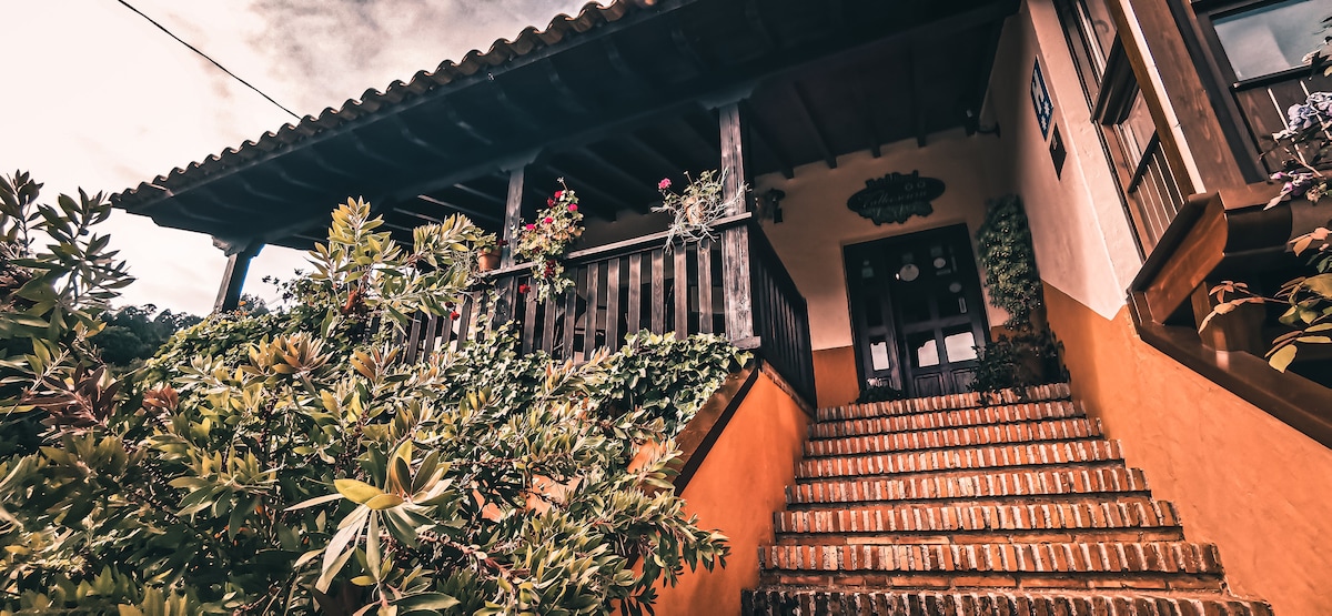 The entrance features a rustic wooden balcony adorned with flowers, leading to a welcoming double door. A set of terracotta steps ascends through lush greenery, providing an inviting pathway to the home, framed by natural elements.