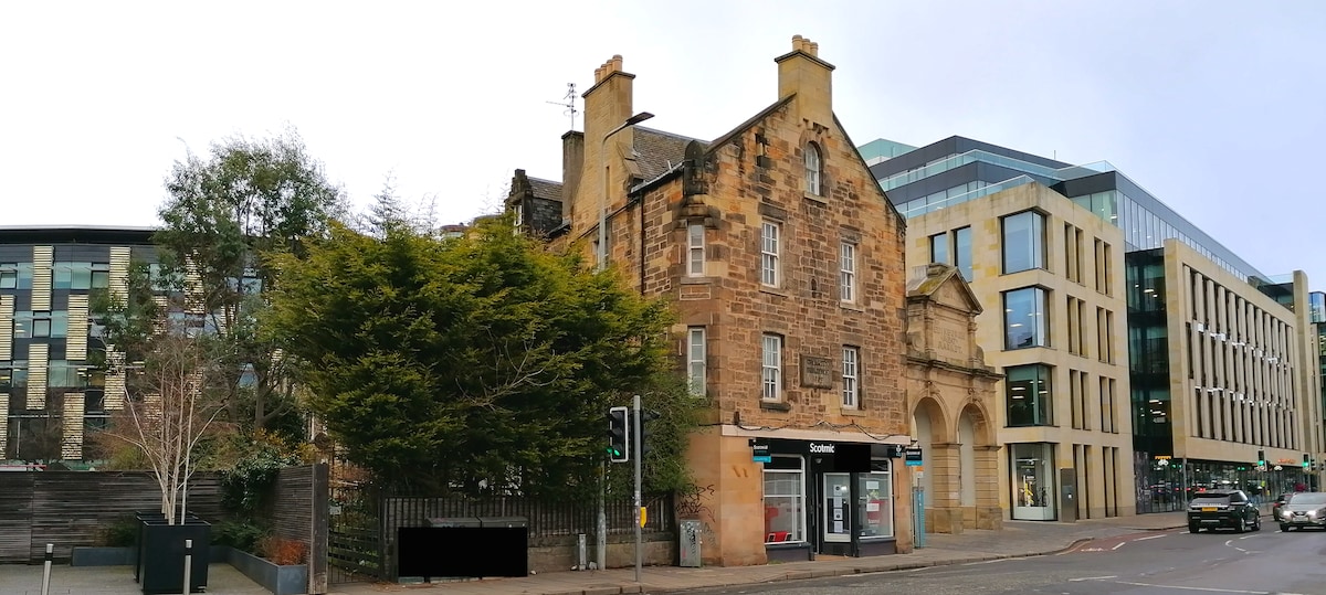 An architectural contrast is presented with an old stone building featuring large windows and a decorative entrance, flanked by modern structures showcasing glass facades. Lush trees provide greenery to the scene, creating a blend of history and contemporary design.