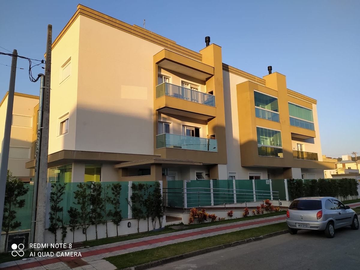 The exterior of the building displays a modern design with multiple balconies framed by glass railings. A well-maintained garden with shrubbery and flowers is situated in front. A driveway runs alongside, accommodating parked vehicles, and ample natural light bathes the façade in the late afternoon.
