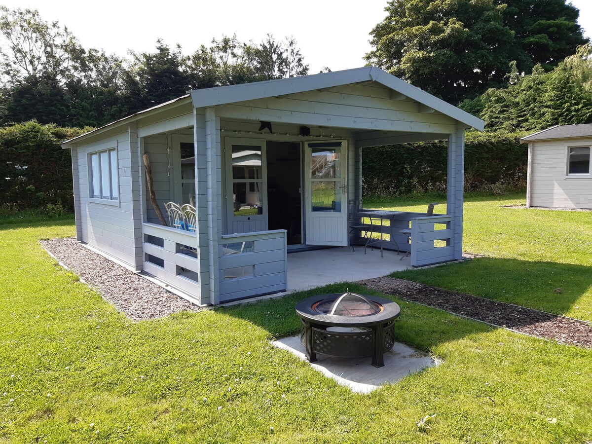 A cabin is presented, featuring a light blue exterior with a covered porch and large windows. Nearby, a fire pit sits on a concrete slab, surrounded by a well-maintained grass area. Outdoor seating is visible, inviting relaxation in a tranquil setting.