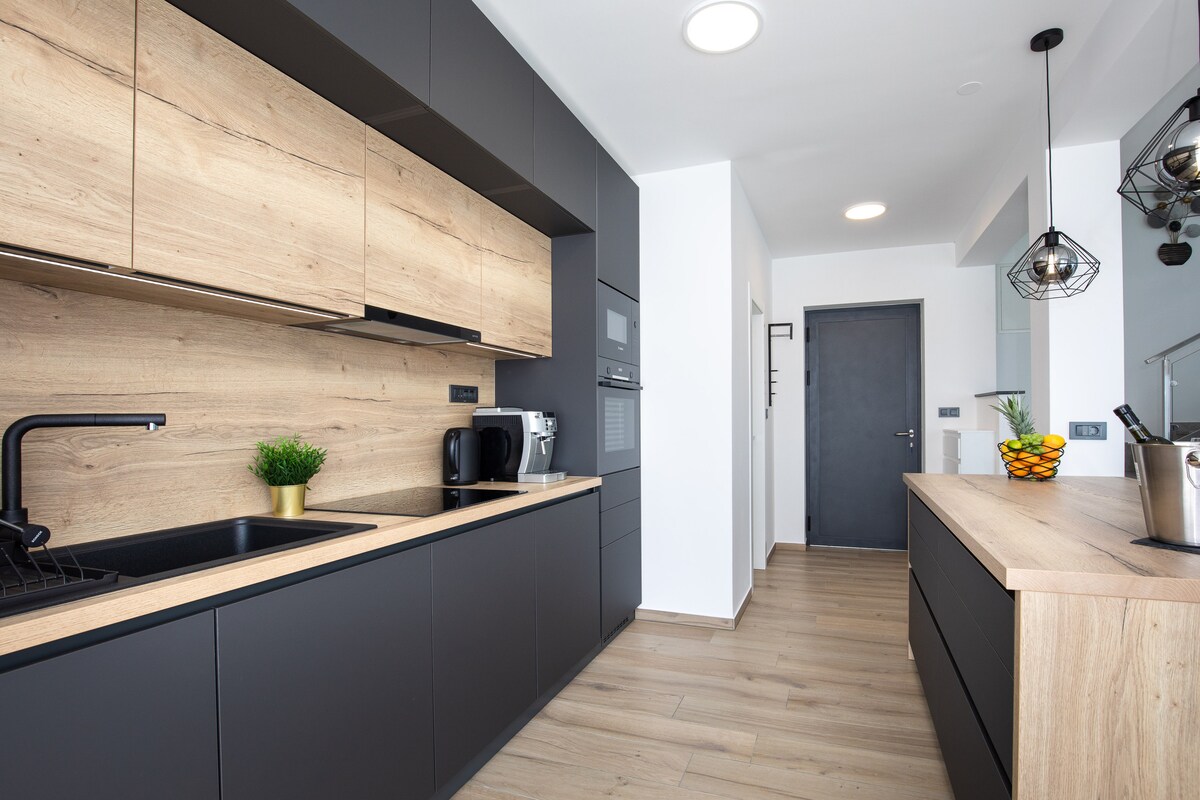 A modern kitchen features sleek cabinetry in dark tones, complemented by warm wooden accents. The space includes a black sink and a variety of appliances, with natural light illuminating the area. A fruit bowl sits on the countertop, adding a touch of color.