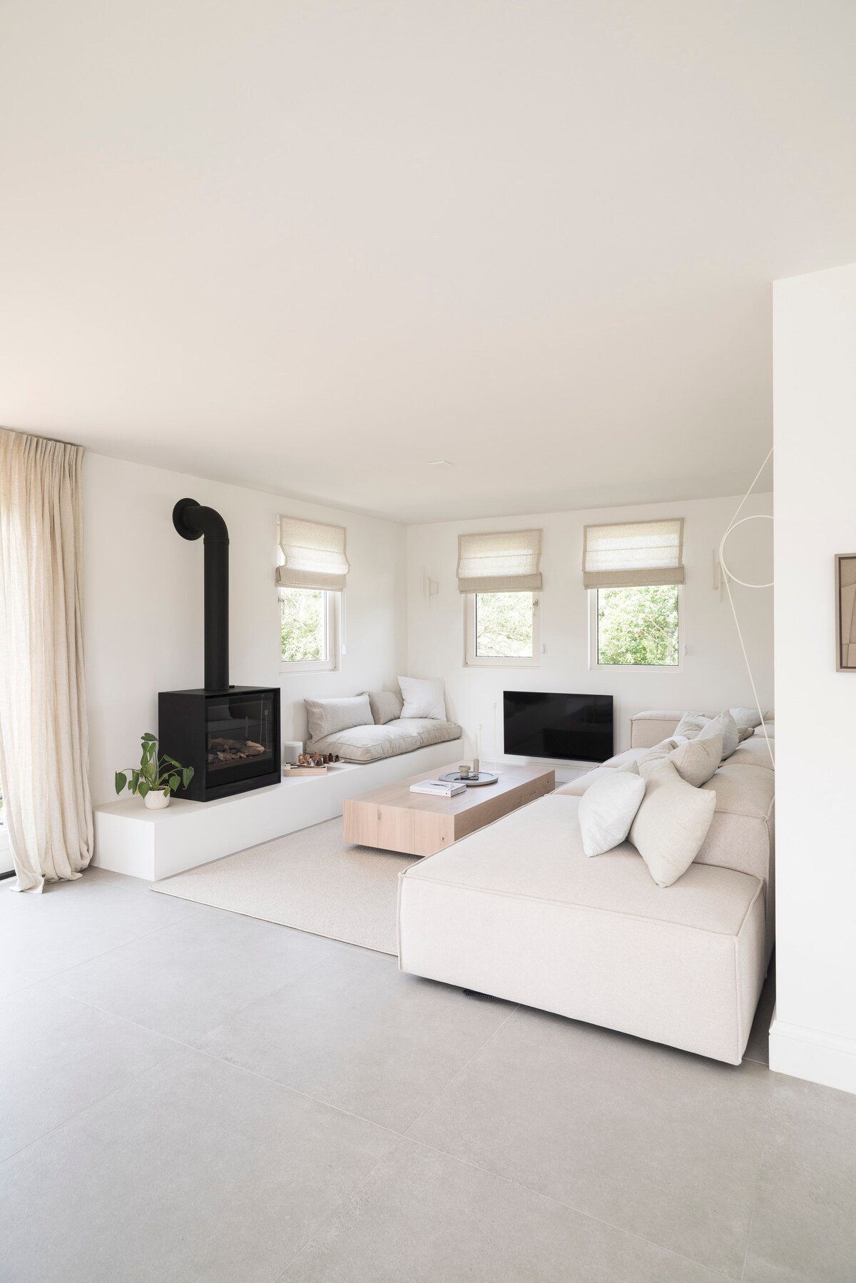 A bright living area features light-colored furniture and a minimalist design. Two spacious sofas are arranged around a central coffee table, with a modern gas fireplace adding warmth. Large windows allow natural light to fill the room, complemented by soft curtains.