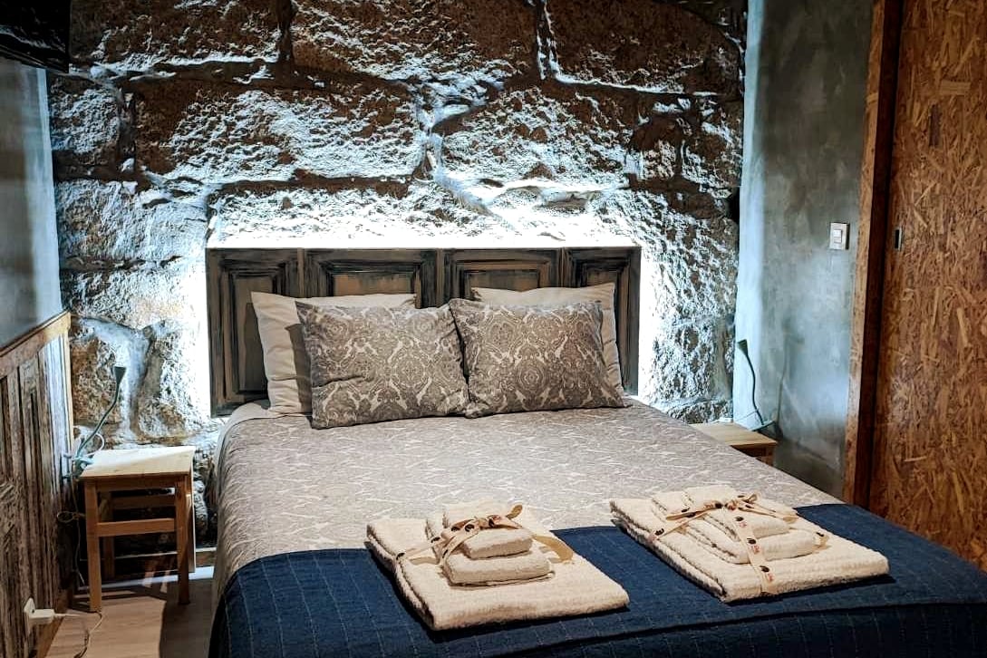 A cozy bedroom features a spacious bed with a patterned coverlet and multiple pillows. Elegant lighting illuminates exposed stone walls, enhancing the rustic appeal. Two small nightstands are positioned on either side of the bed, each topped with decorative items.