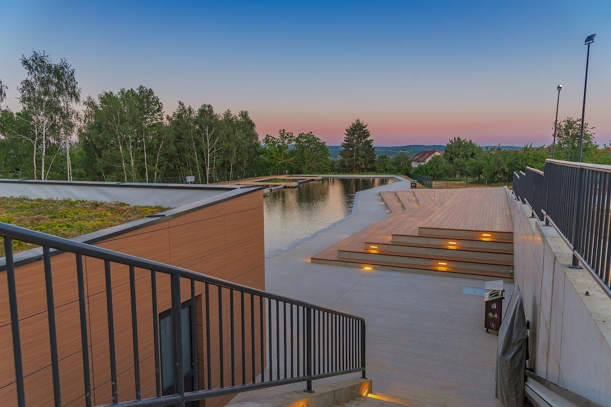 A serene outdoor space is presented, featuring a calm water body reflecting the evening sky. Steps lead down from a wooden platform, with landscaped greenery visible at the edges. Soft lighting enhances the pathway along the water, creating an inviting atmosphere for relaxation.