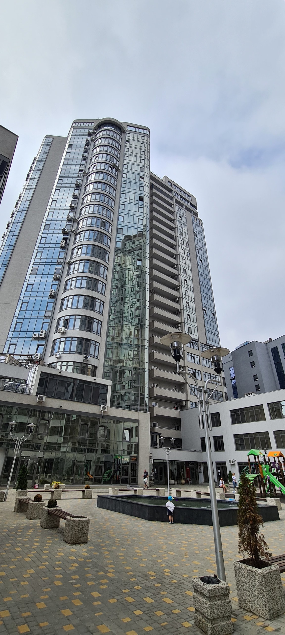 The modern apartment complex features a sleek glass facade, showcasing multiple stories. A calm courtyard with paved walkways and decorative planters is visible in the foreground, while residents can be seen enjoying the outdoor space.