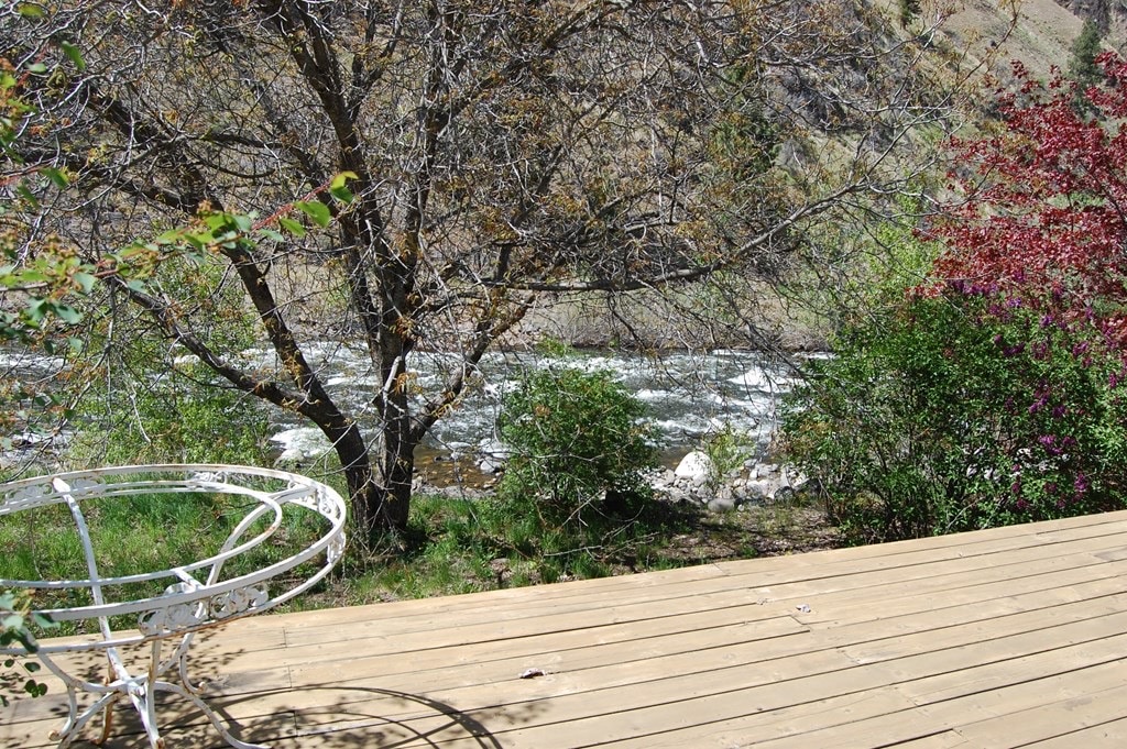 A wooden deck extends towards a gently flowing river, framed by lush green grass and blooming bushes. A white metal chair is positioned nearby, offering a view of the water. Trees provide shade and texture to the picturesque outdoor setting.