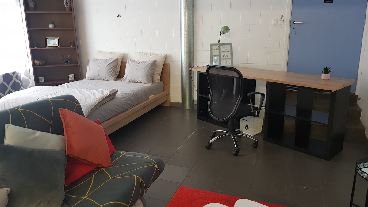 A studio room features a spacious bed with light bedding against a wall. A desk and chair are positioned next to a wall shelf displaying decorative items. A lounging chair adorned with colorful cushions adds comfort to the setting. Natural light is invited through a nearby window.