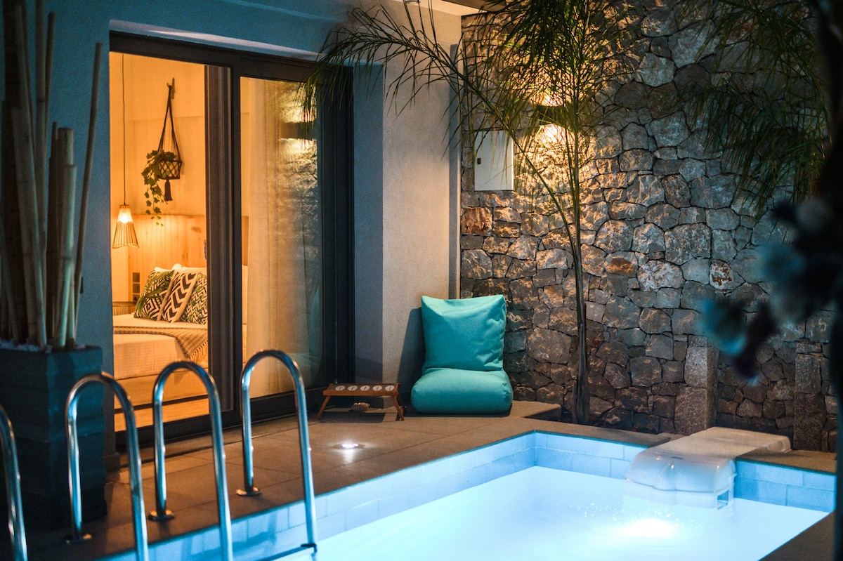 A serene outdoor space is highlighted by a small swimming pool with soft blue lighting. A cozy seating area is visible beside the pool, adorned with a bright cushion. The stone wall behind enhances the ambiance, complemented by gentle illumination from wall sconces.