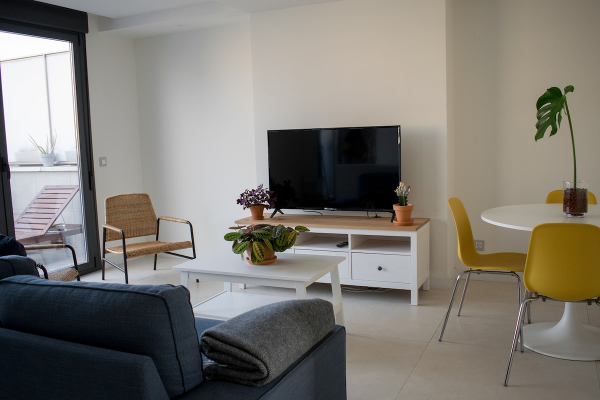 A modern living area features a flat-screen television mounted on a light-colored console. A round dining table with yellow chairs is positioned next to a potted plant. A soft gray blanket drapes over a blue sofa, creating a relaxed setting near sliding glass doors.