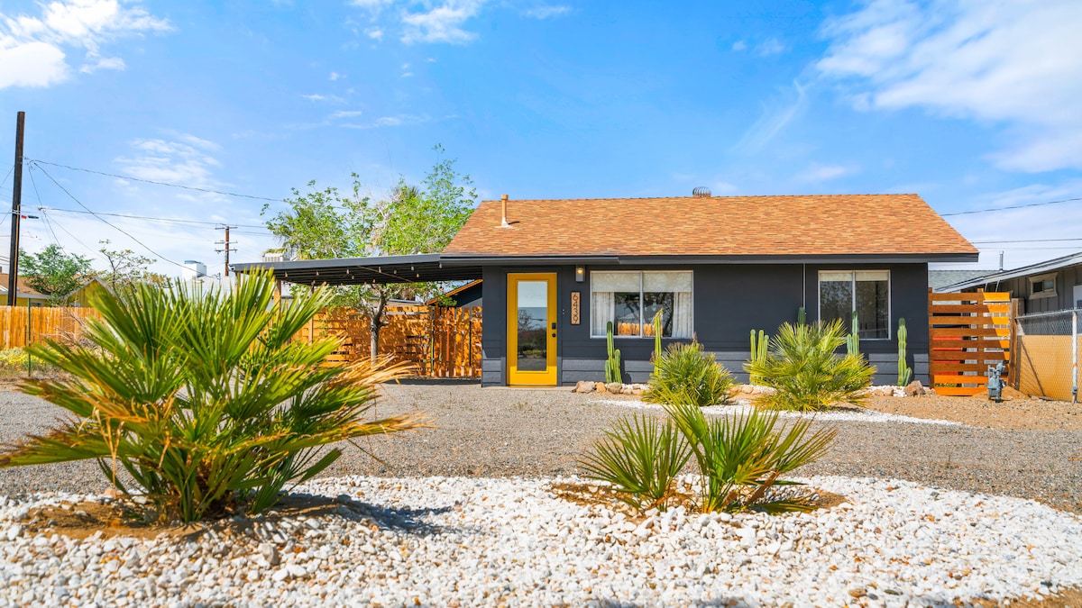 The exterior of the modern retreat is showcased with a well-manicured front yard featuring a mix of desert plants and pebbled pathways. The home’s muted facade is accented by a vibrant yellow door and a covered carport. Clear blue skies complement the inviting atmosphere.