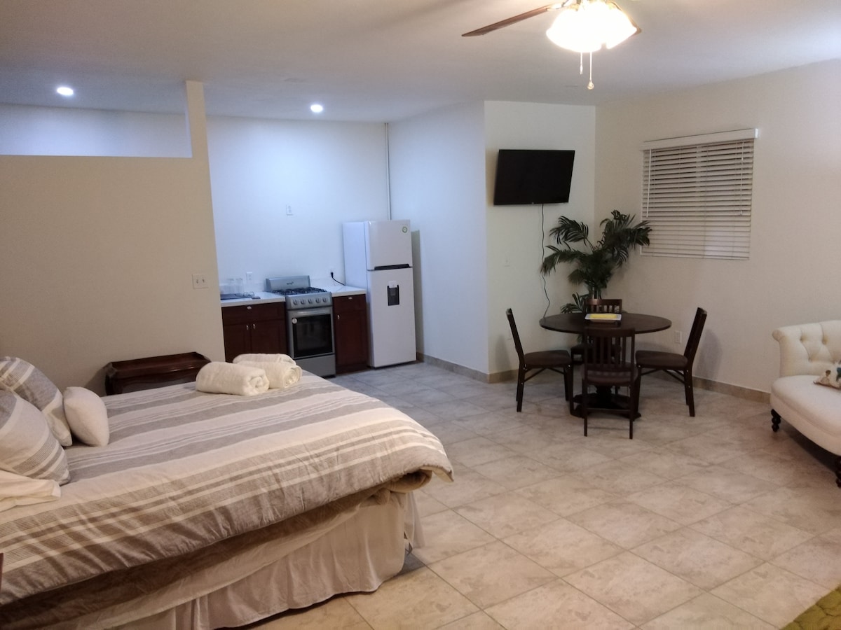 A studio apartment is shown, featuring a spacious bed with striped bedding and plush pillows. A small dining table is present, paired with chairs. A kitchenette with a refrigerator and stove is located in the corner, and a wall-mounted TV is visible. Natural light enters through a window.