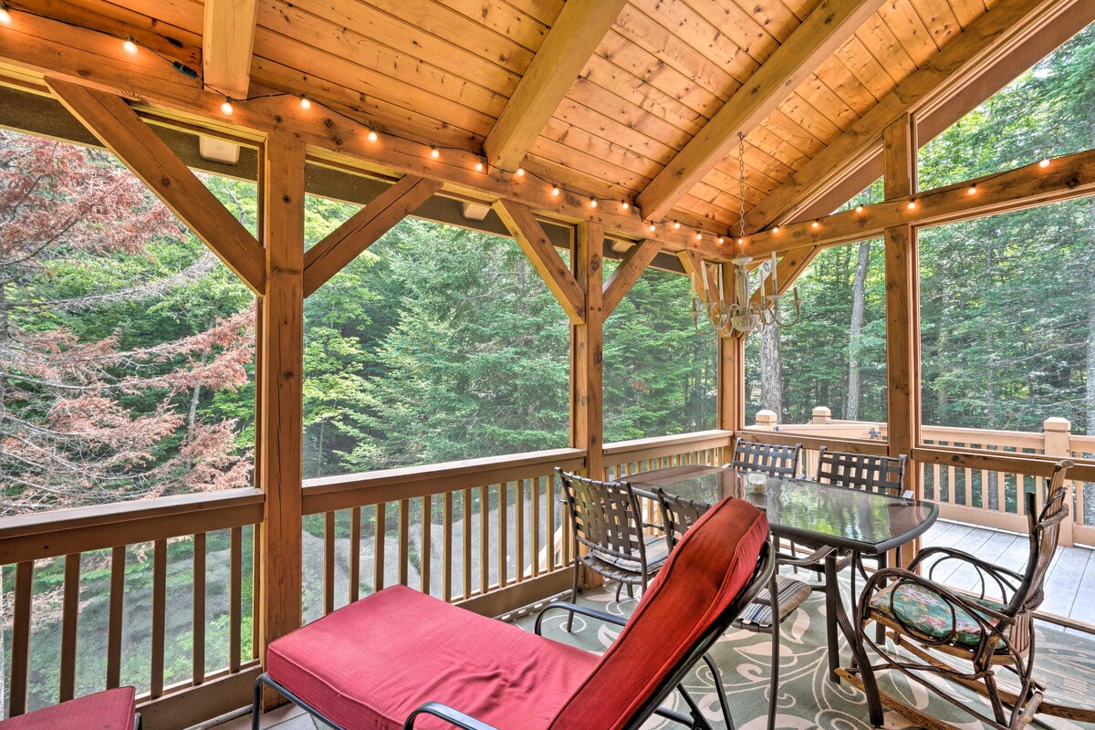 A screened-in porch features wood beams and a vaulted ceiling, illuminated by soft string lights. Outdoor furniture includes a dining table with chairs and a lounge chair, all positioned to enjoy views of the surrounding forest. A patterned rug adds comfort to the space.