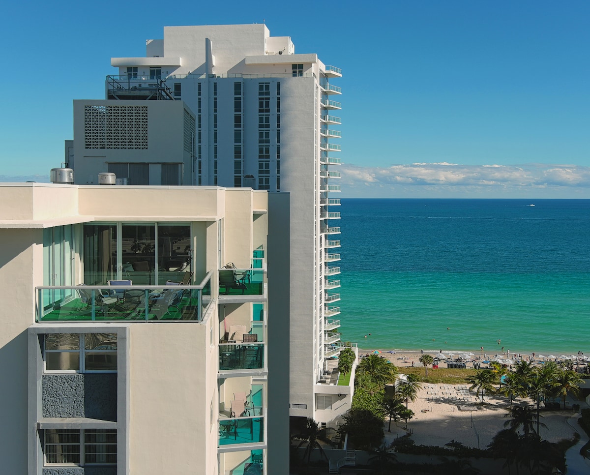 An exterior view of the Sian Condominium showcases its modern design against a backdrop of the ocean. The turquoise water meets the beach, while people can be seen enjoying the sun. The building features balconies, offering relaxing outdoor spaces.