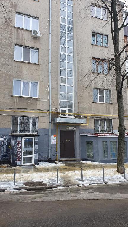 The exterior of a multi-story building is displayed, featuring a central glass-paneled entrance and narrow horizontal windows. Snow is visible on the ground, and a simple railing runs along the pathway. The facade is characterized by a muted color palette, revealing a blend of gray tones.