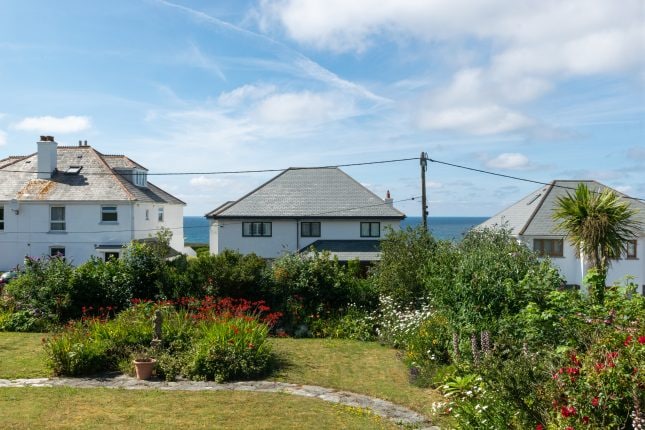 A view of adjacent houses is captured, showcasing their white exteriors against a clear sky. Lush greenery and colorful flowers fill the foreground garden, while the ocean is visible in the distance, complementing the tranquil coastal setting.