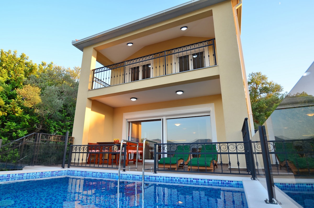 A two-story villa is seen from the poolside, featuring a spacious terrace with outdoor dining furniture. The balcony above offers additional seating. Large glass doors lead to the interior, enhancing the connection between indoor and outdoor living spaces, surrounded by lush greenery.