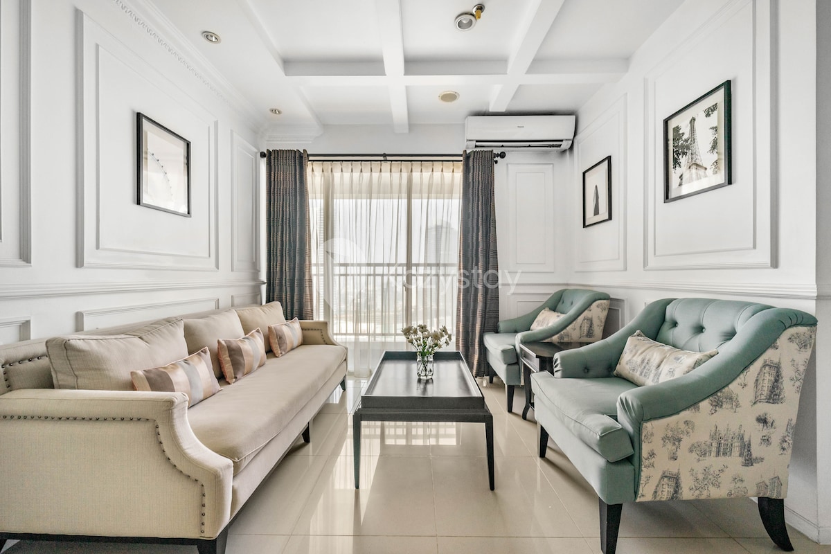 The living area features a neutral-toned sofa alongside two accent chairs in a soothing shade. A low coffee table sits at the center, complemented by floral decor. Large windows allow natural light to enter, while sheer curtains add a layer of softness to the space.