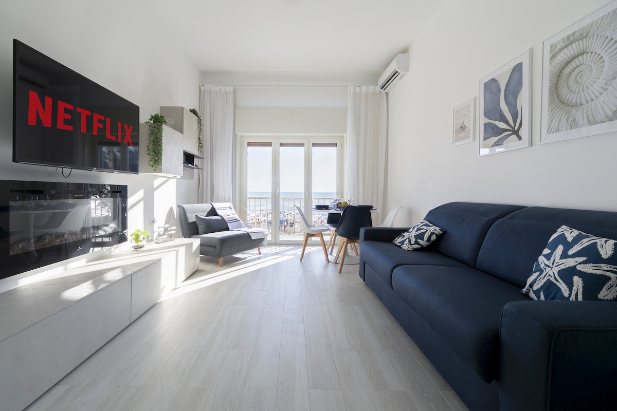 A bright and airy living area features large windows overlooking the sea. A comfortable dark blue sofa and a light gray chair are arranged around a coffee table. A sleek entertainment unit houses a TV, while light-colored artwork adorns the walls.