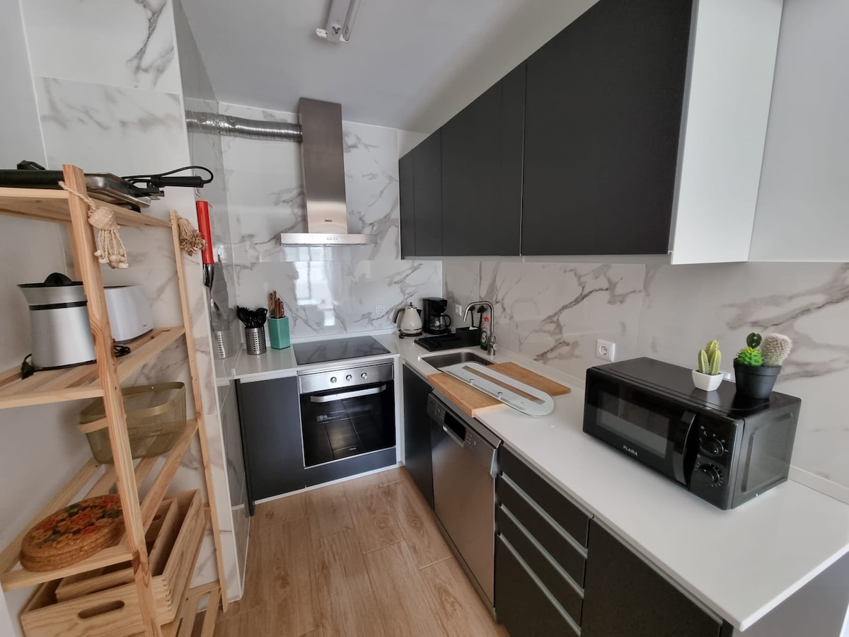 A modern kitchenette is equipped with a sleek design, featuring black cabinetry and white marble backsplash. Appliances include a built-in oven, microwave, and kettle, with utensils arranged nearby. A wooden shelving unit holds cookware, contributing to the functional layout.