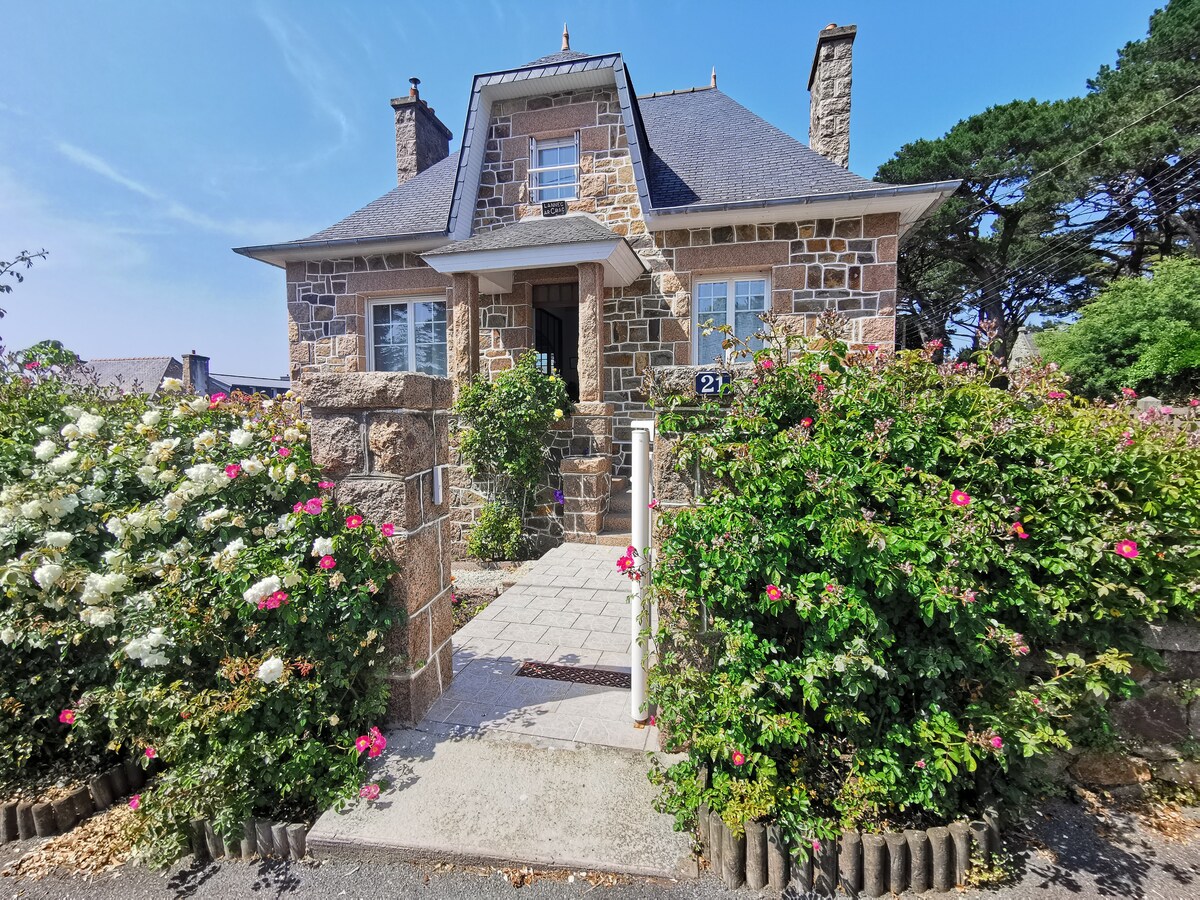 A charming stone house is showcased, framed by vibrant flowering bushes. The front entrance features a small pathway leading to a welcoming door, with a sloped roof and neatly placed stone accents enhancing its rustic appeal. Lush greenery surrounds the property, reflecting a tranquil atmosphere.