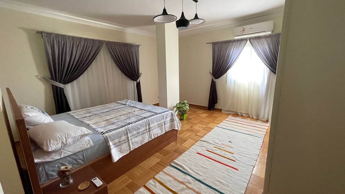 A spacious bedroom features a comfortable bed with neatly arranged bedding. Large windows are adorned with flowing curtains, enhancing natural light. A decorative rug with colorful stripes is placed on the wooden flooring, and a small plant adds a touch of greenery.