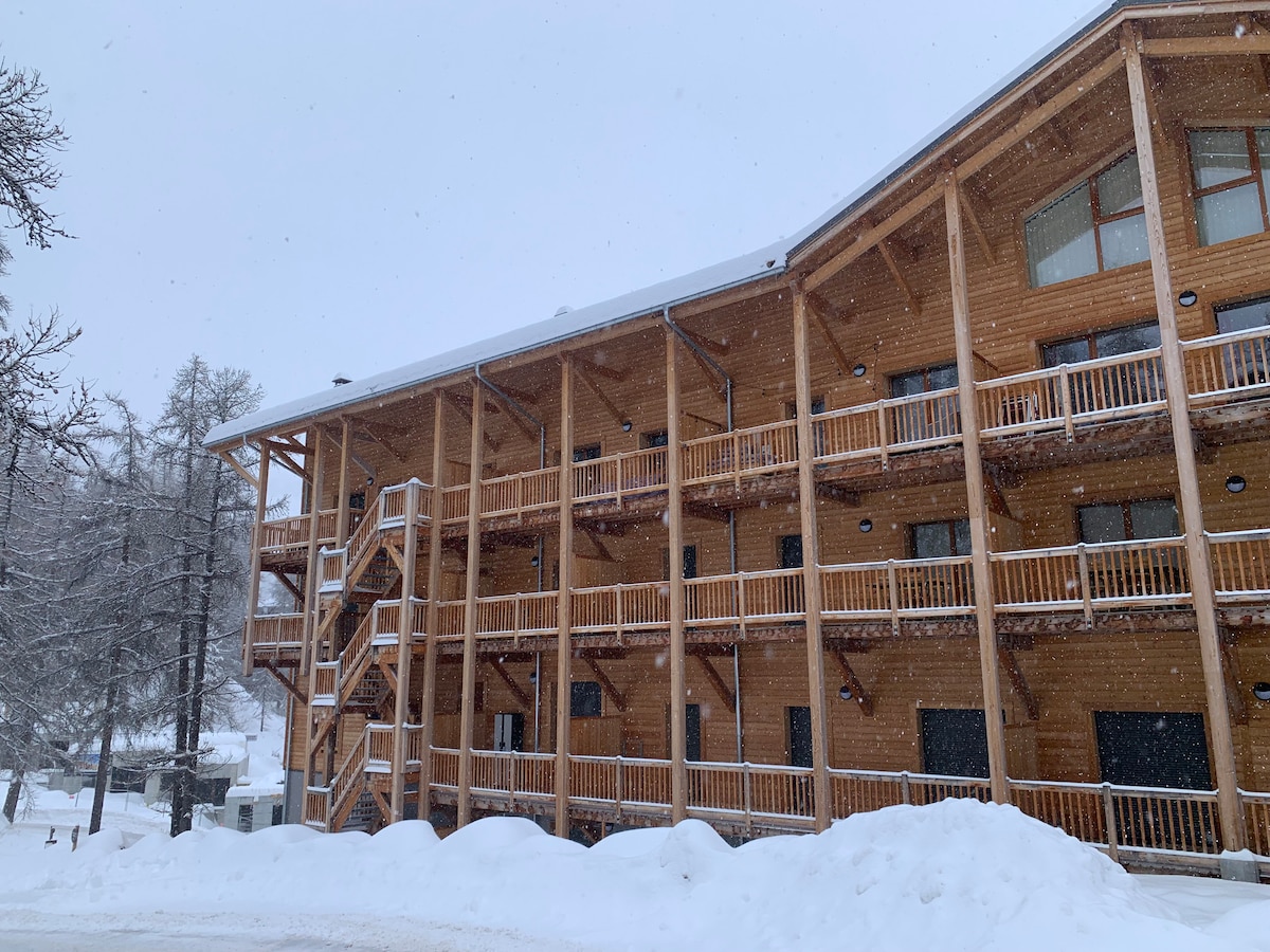 The exterior of a wooden chalet-style building is visible, featuring multiple balconies and a symmetrical design. Snow falls gently, covering the ground and adding to the winter atmosphere. The warm wooden finish contrasts with the white snow, creating a cozy appearance.