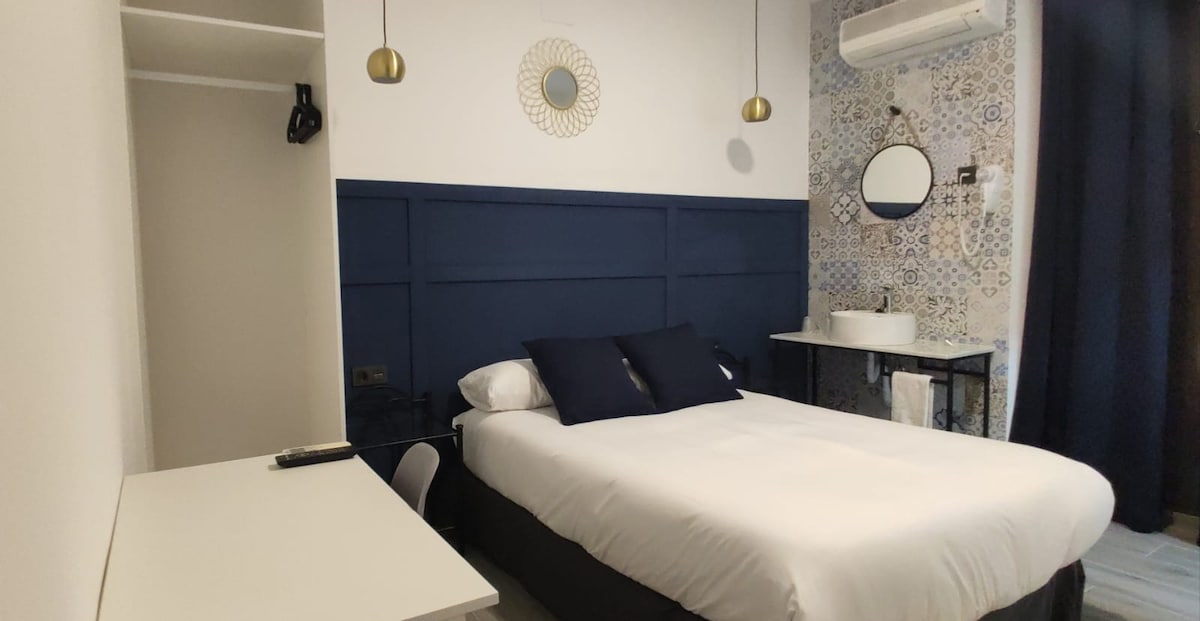 A modern sleeping area features a spacious bed with white linens and dark pillows. A wall-mounted mirror and a round decorative piece are visible above a black side table. Air conditioning is installed, and a neatly arranged workspace is located to the left.