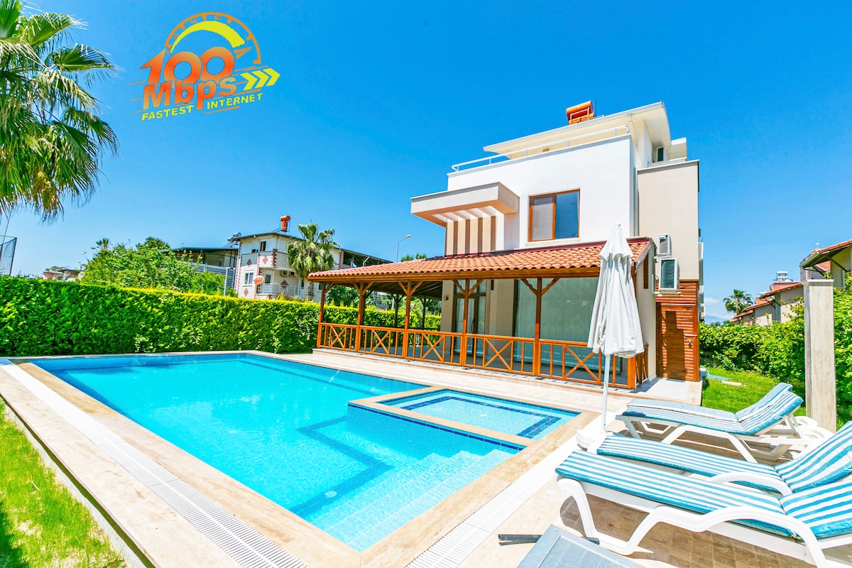 A modern villa is featured, showcasing a crystal-clear pool surrounded by comfortable lounge chairs. A shaded gazebo provides relaxation space, complemented by lush greenery that surrounds the property. The structure displays clean lines and a welcoming atmosphere under the clear blue sky.
