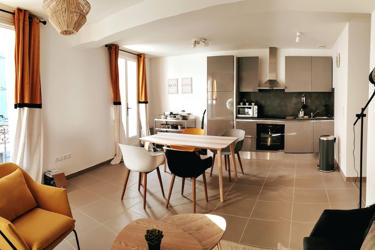 A modern open-plan kitchen and dining area features a mix of seating options, including two yellow-accented chairs. Large windows allow natural light to illuminate the space. The kitchen is fully equipped with sleek cabinetry and appliances, all set against neutral-toned walls and tiled flooring.