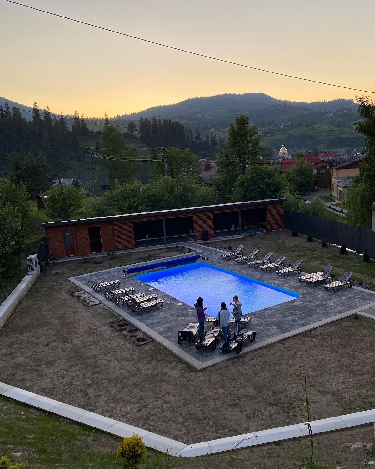 An outdoor area features a well-designed pool surrounded by tiled decking and multiple lounge chairs. The setting is complemented by scenic mountain views and a backdrop of trees at sunset, creating an inviting atmosphere for relaxation.
