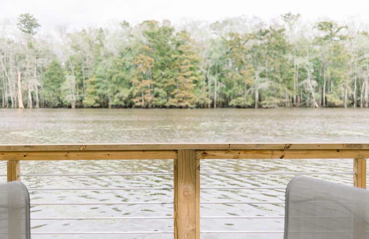 A wooden railing provides a view of calm waters reflecting the surrounding trees. The natural scenery features lush greenery on the opposite bank, creating a serene backdrop. The water's surface is gently rippled, enhancing the tranquil atmosphere.