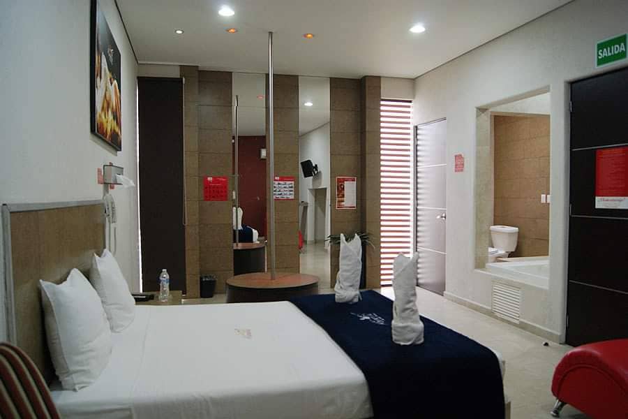 A modern room features a spacious bed with white linens and decorative pillows. A small round table and striped chair are positioned nearby. A shower area is visible, alongside a separate bathroom. Soft lighting from ceiling fixtures creates an inviting environment.