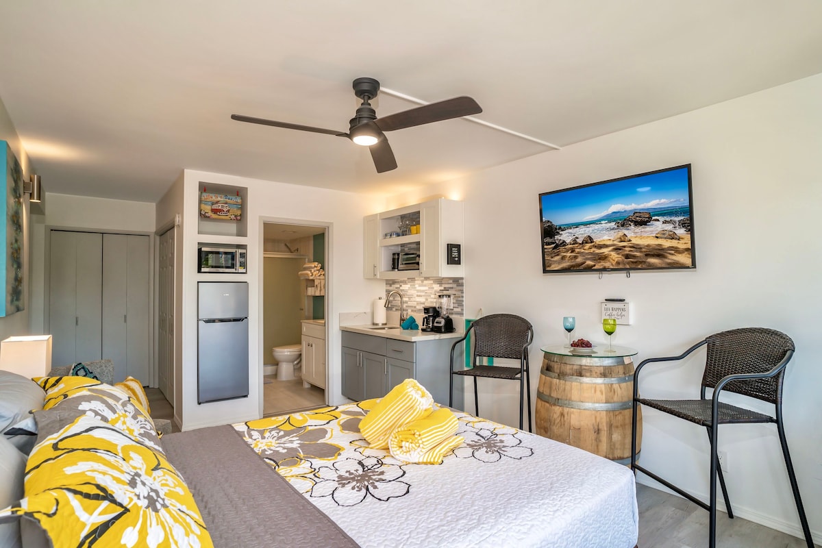 A modern studio space is showcased with a spacious bed layered with decorative yellow and gray linens. A kitchenette with a microwave and refrigerator is present, along with a flat-screen TV mounted on the wall. Outdoor seating is arranged beside a barrel table.
