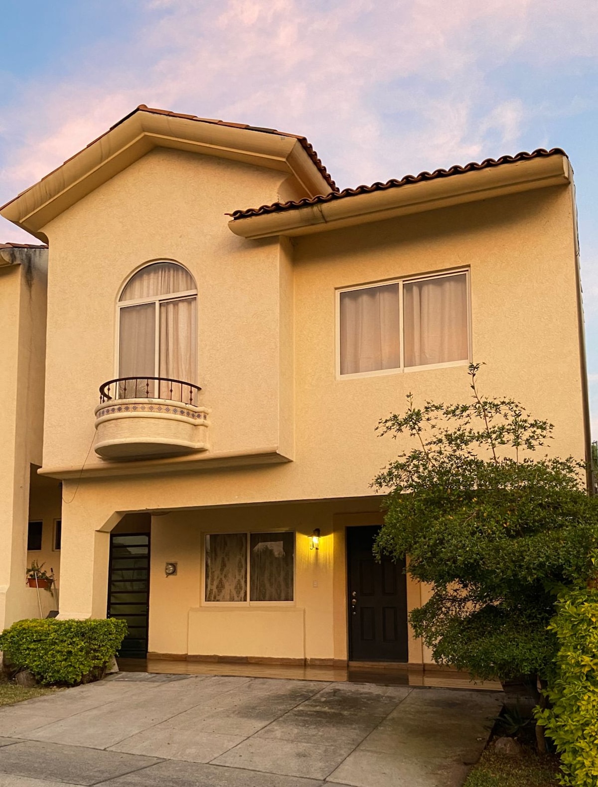 The front of a two-story house features a unique architectural design with an arched window and a small balcony above the entrance. Lush greenery surrounds the entrance, complemented by a well-maintained pathway leading to the door. Soft lighting highlights the entrance during dusk.