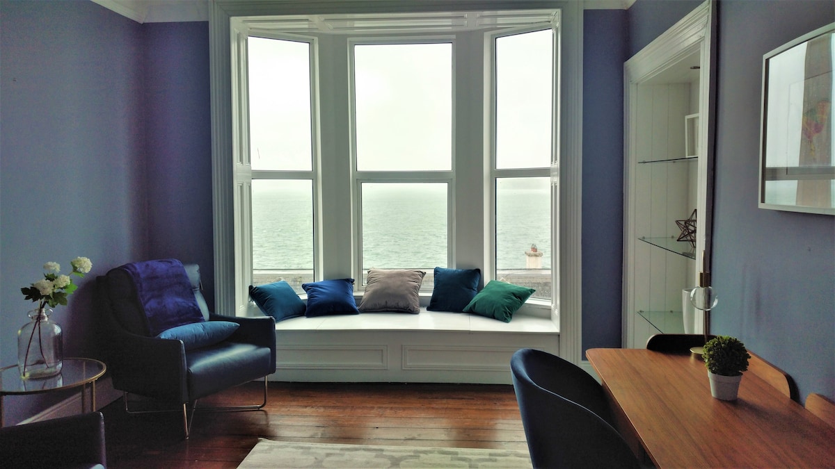 A bright seating area features a window bay with cushions in various colors, offering a view of the sea. Blue walls enhance the space, complemented by a wooden dining table and chairs nearby. Natural light fills the room, creating an inviting environment.