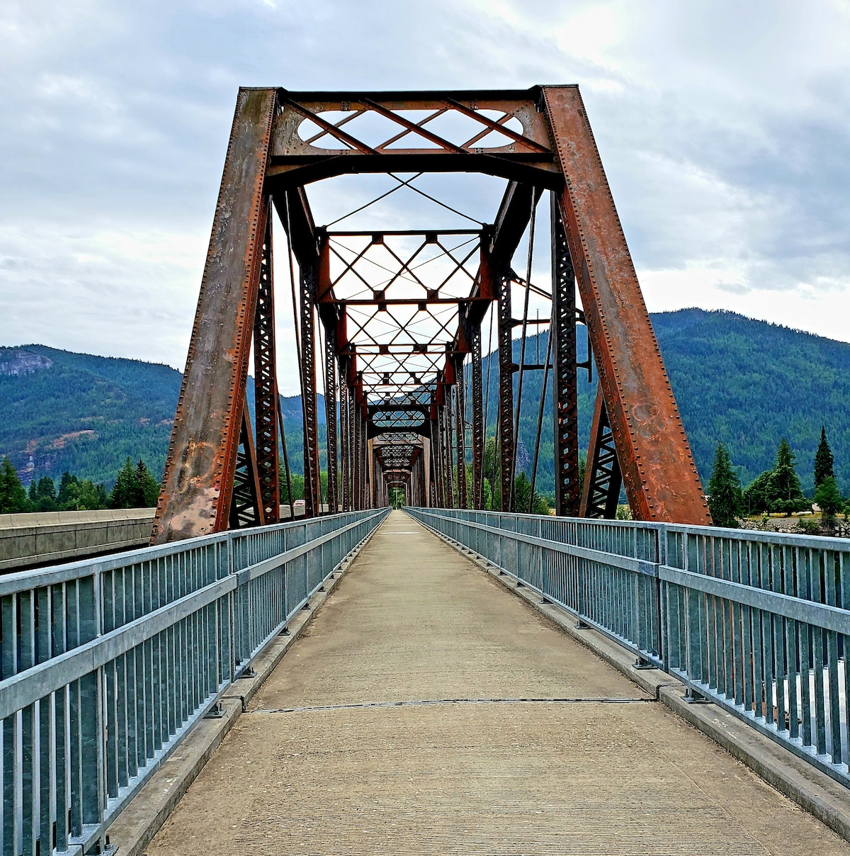 An old metal bridge spans the river, with weathered red beams and a concrete walkway. Lush green mountains rise in the background under a lightly clouded sky. The structure creates a sense of journey across the water.
