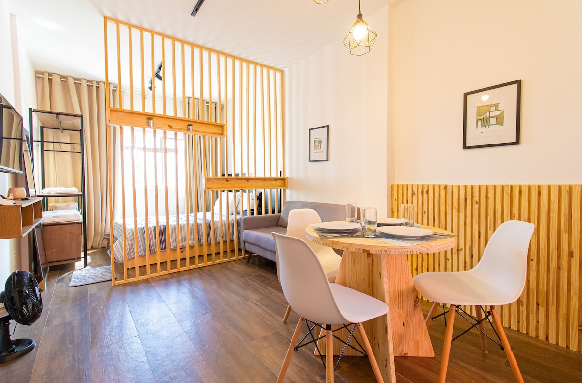 An open living space is divided by wooden slats, revealing a cozy sleeping area with a spacious bed. A circular dining table with three chairs is set in the foreground, and a small desk with a TV is positioned nearby, enhancing usability in the apartment.