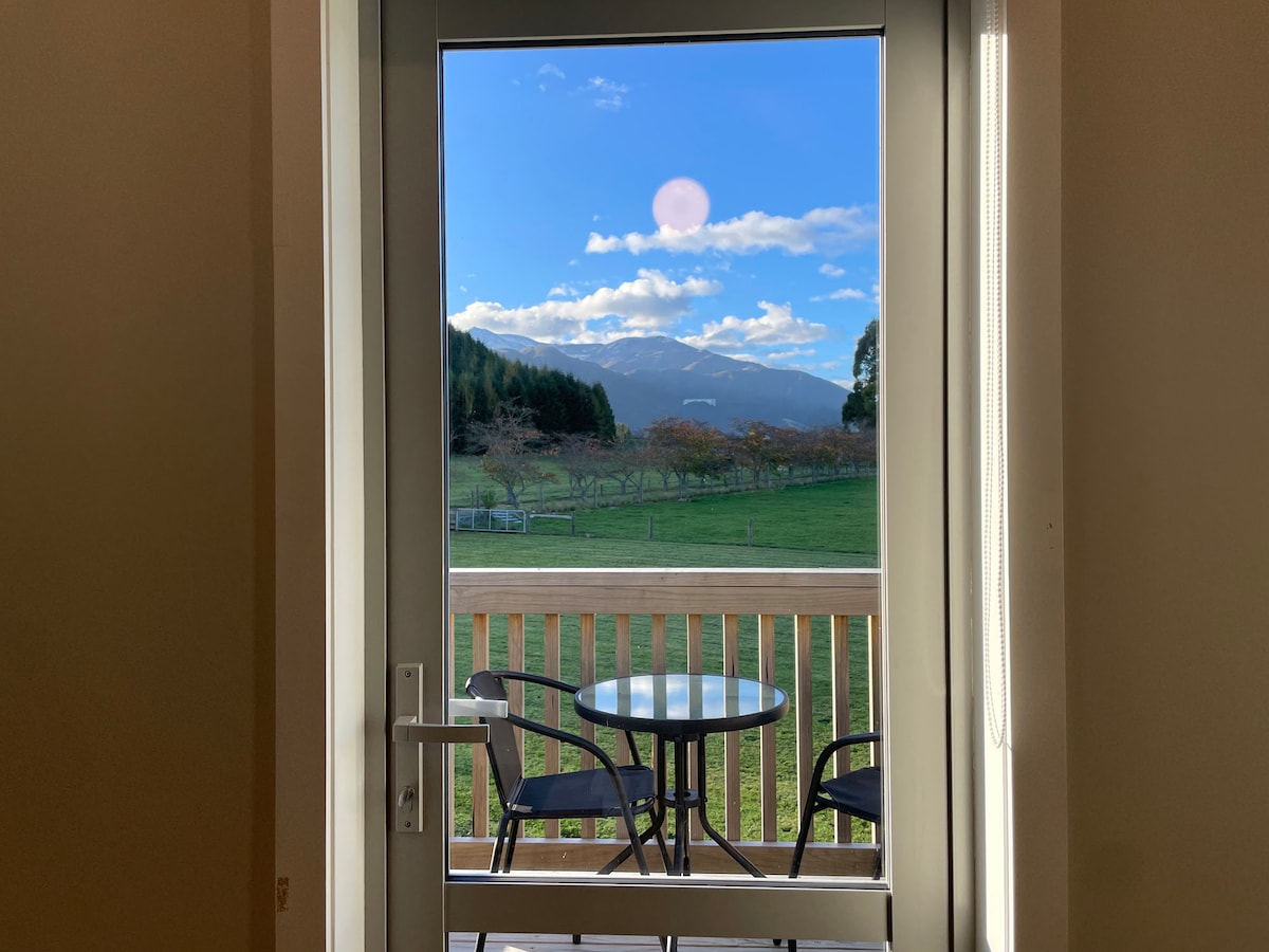 A door opens to reveal a private balcony featuring a round table and two chairs. Beyond, expansive views of the Southern Alps are visible under a blue sky, with lush fields framed by distant mountains and soft clouds.