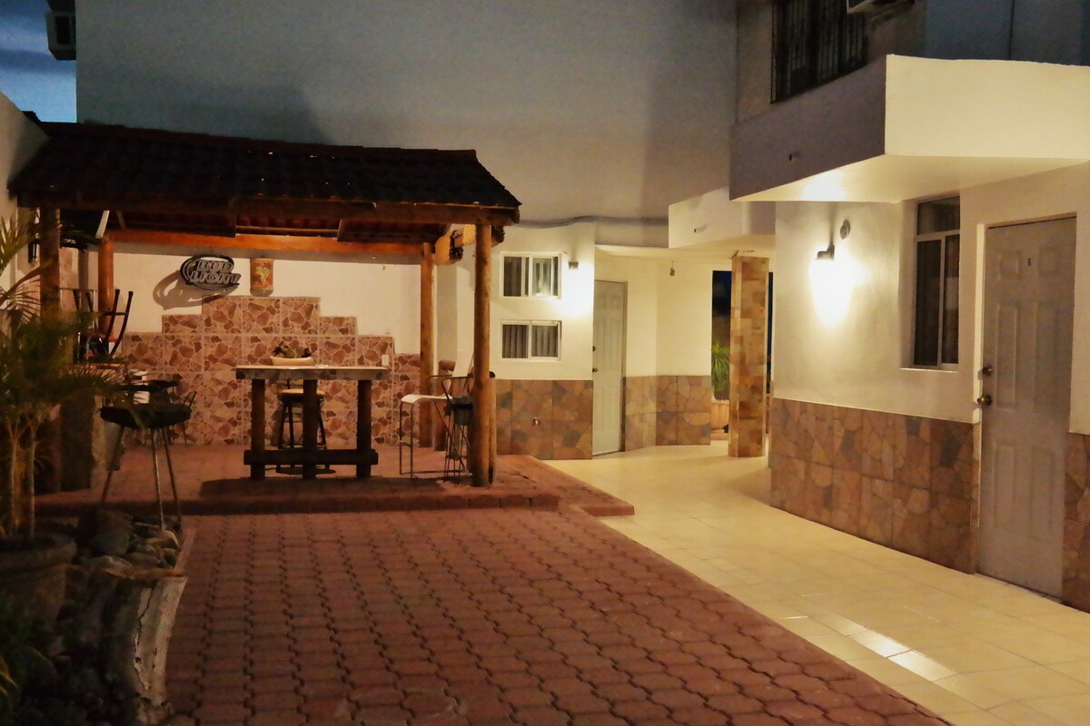 An outdoor area features a covered seating arrangement with a table and chairs, set against a stone wall. Pathways lined with tiles lead to various entrances, illuminated by soft lighting. The surroundings are enhanced by decorative plants, creating a peaceful and functional space.