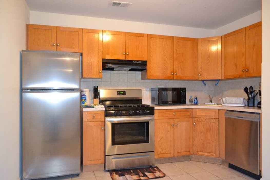 The kitchen features light wooden cabinetry and stainless steel appliances, including a refrigerator and oven. A microwave is placed on the countertop, alongside various kitchen utensils. Neutral tile flooring complements the warm tones of the cabinets, creating a functional cooking space.