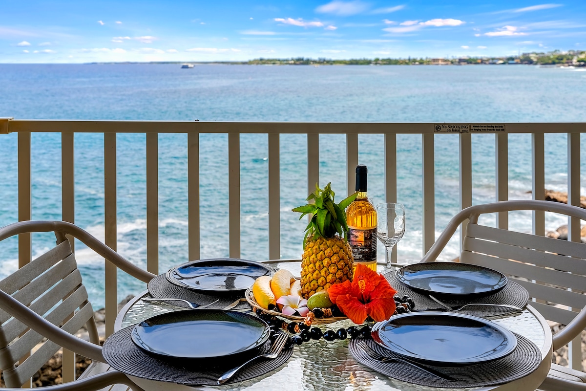 A dining table set for two overlooks the ocean, featuring black plates and wine glasses. A centerpiece showcases a pineapple surrounded by tropical fruits, flowers, and greenery. The sparkling water and distant coastline create a scenic backdrop.