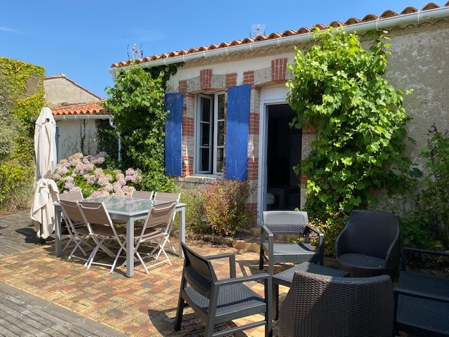 An outdoor terrace is presented with a dining table surrounded by chairs, complemented by lounge seating. Lush greenery and flowering plants provide a natural backdrop. Bright blue shutters frame the doorway, allowing light to fill the interior space. A sun umbrella offers shade.