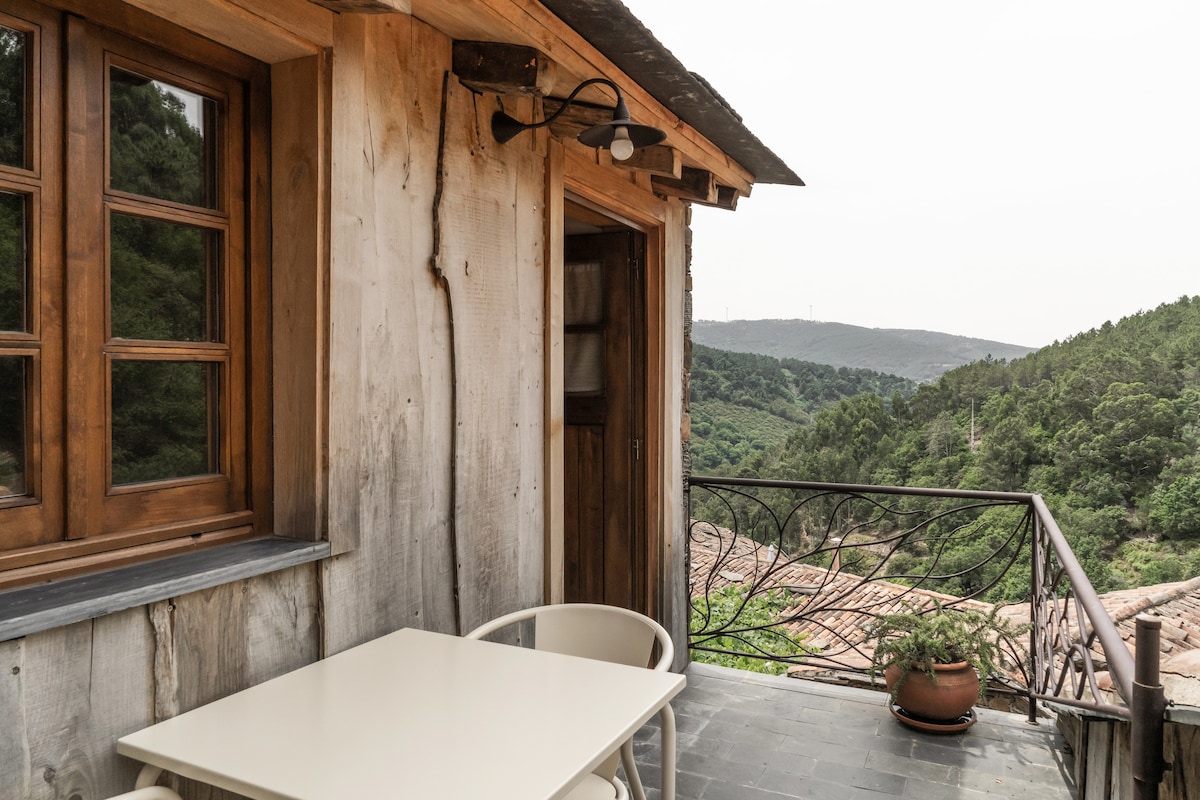 A small balcony features a light-colored table and two chairs, surrounded by a wooden railing. Green hills extend into the distance, providing a natural backdrop. The door leading to the interior is made of wood, complementing the rustic aesthetic of the space.