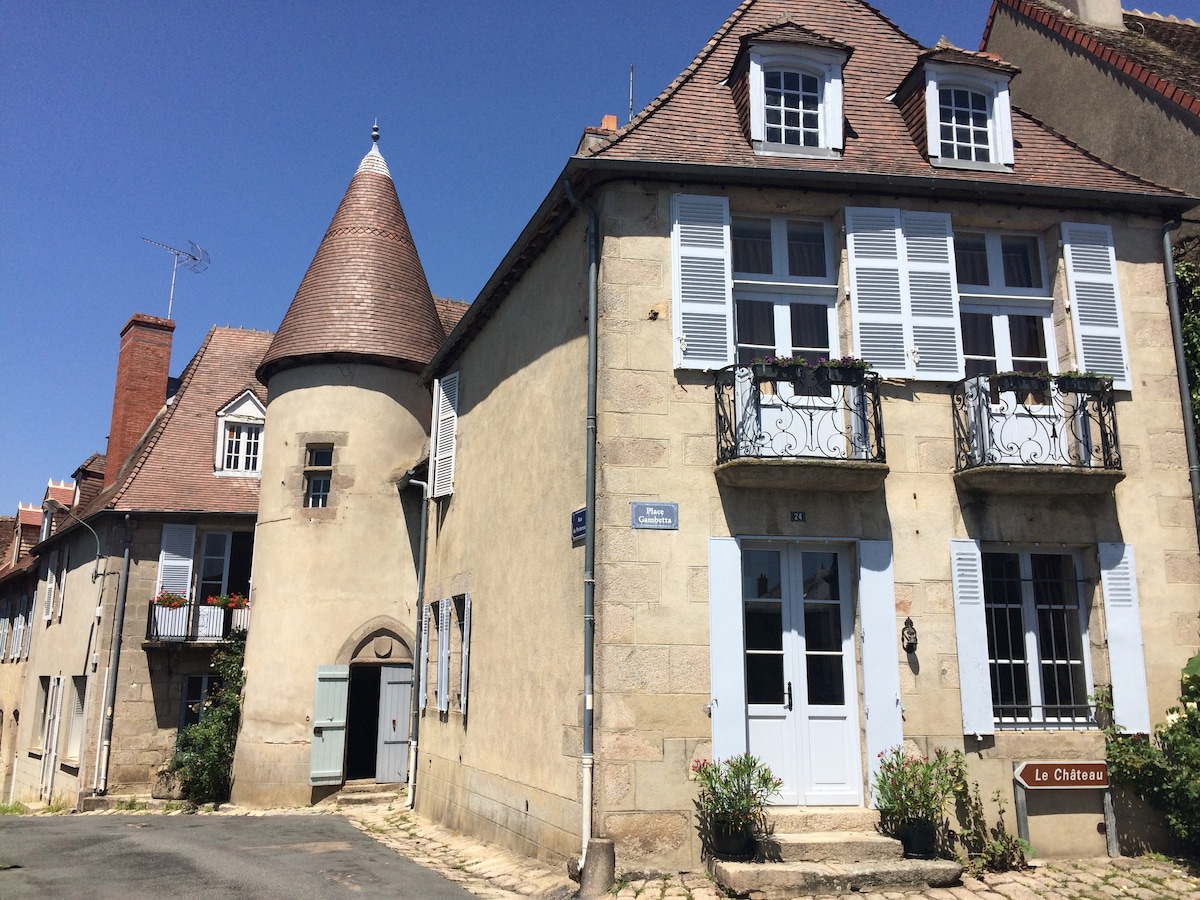 The exterior of a beautifully restored historical building features a charming turret and multiple windows adorned with white shutters. A welcoming blue door is prominently displayed, complemented by subtle landscaping around the entrance, reflecting the building's architectural heritage.