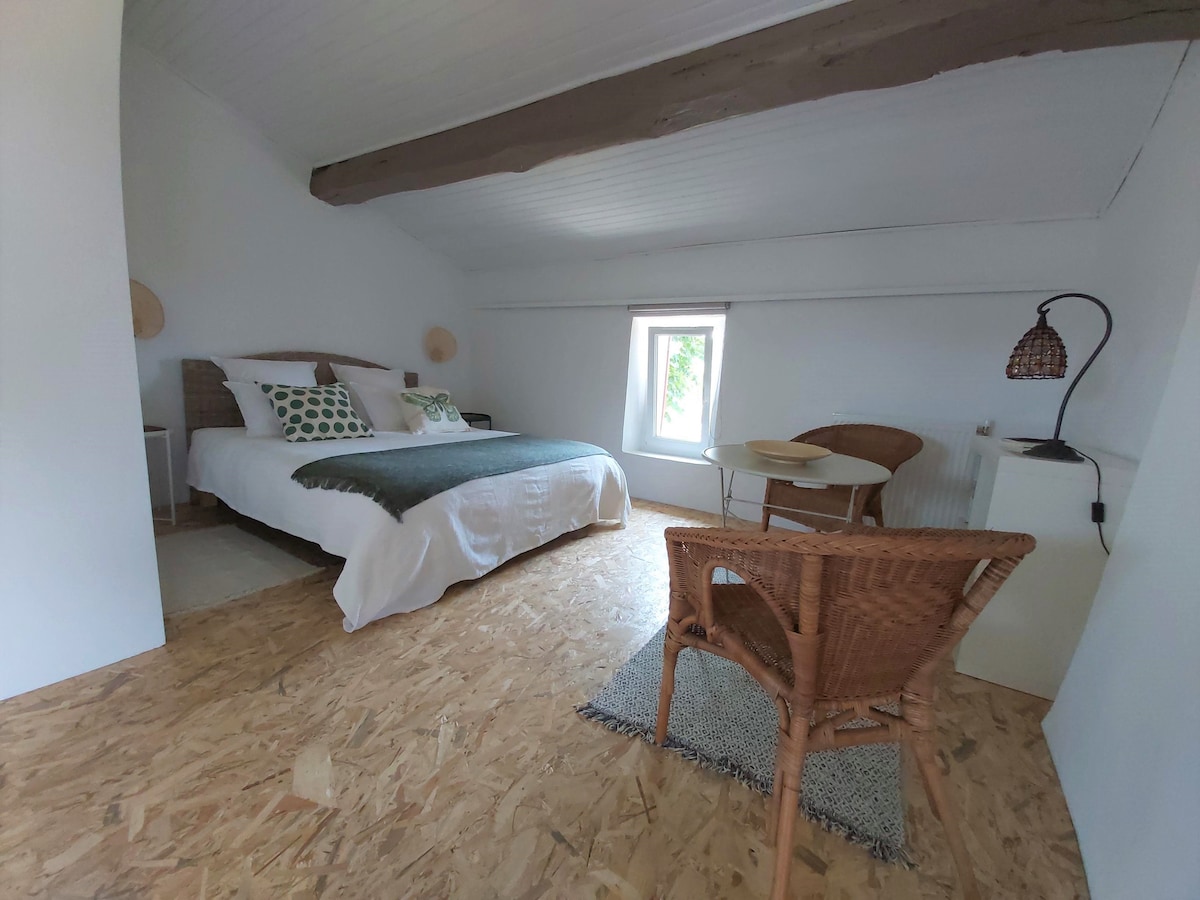The studio features a spacious bed with light-coloured bedding and decorative pillows. A small table and two chairs are arranged nearby, while a window allows natural light to enter. The floor is covered with wooden panels, offering a rustic look.