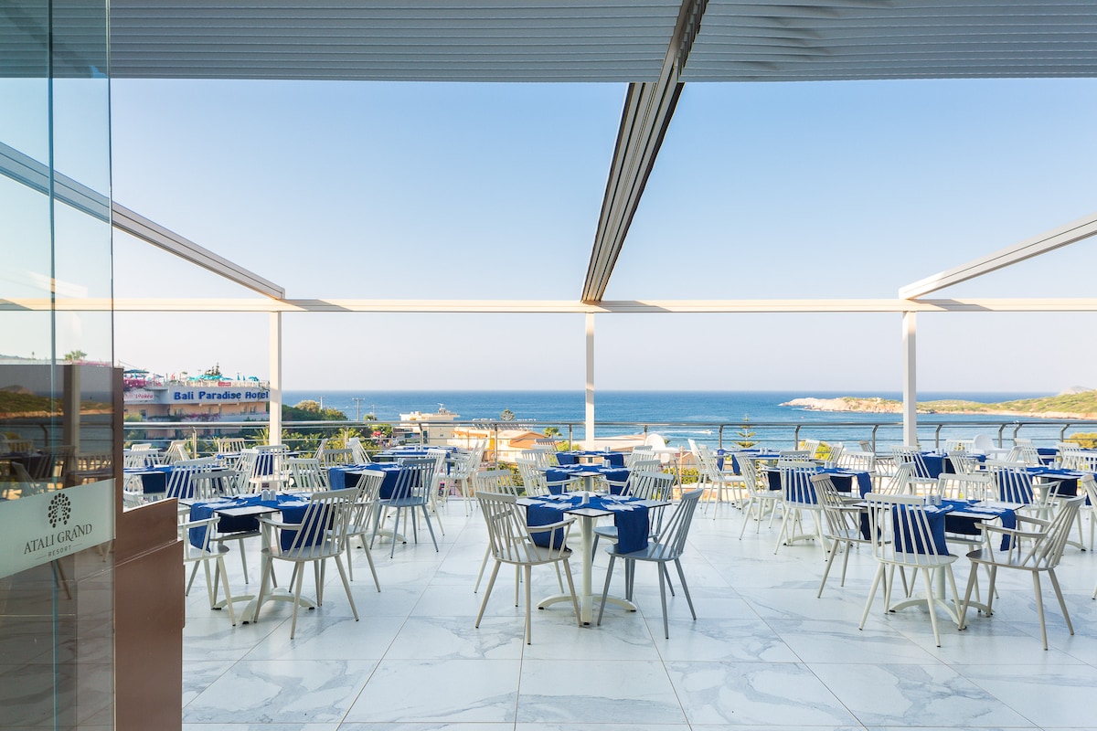 An open-air restaurant features modern dining furniture arranged on a marble terrace, offering views of the sea. The space is complemented by large glass walls and an awning, providing shade while allowing natural light to fill the area.